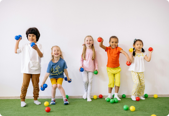 diverse group of 4 to 5 year old children lined against wall playing dodgeball with small colorful plastic balls